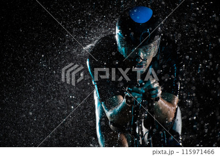 A triathlete braving the rain as he cycles through the night, preparing himself for the upcoming marathon. The blurred raindrops in the foreground and the dark, moody atmosphere in the background add A triathlete braving the rain as he cycles through the night, preparing himself for the upcoming marathon. The blurred raindrops in the foreground and the dark, moody atmosphere in the background add 115971466