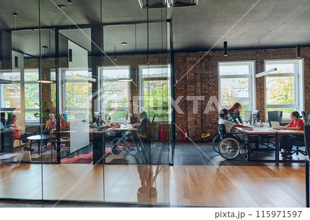 A young business group, including an African American businessman in a wheelchair, collaborates within a modern glass office, actively engaged around a computer and laptop, collectively solving A young business group, including an African American businessman in a wheelchair, collaborates within a modern glass office, actively engaged around a computer and laptop, collectively solving 115971597