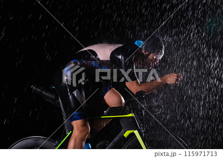 A triathlete braving the rain as he cycles through the night, preparing himself for the upcoming marathon. The blurred raindrops in the foreground and the dark, moody atmosphere in the background add 115971713
