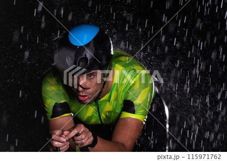 A triathlete braving the rain as he cycles through the night, preparing himself for the upcoming marathon. The blurred raindrops in the foreground and the dark, moody atmosphere in the background add A triathlete braving the rain as he cycles through the night, preparing himself for the upcoming marathon. The blurred raindrops in the foreground and the dark, moody atmosphere in the background add 115971762