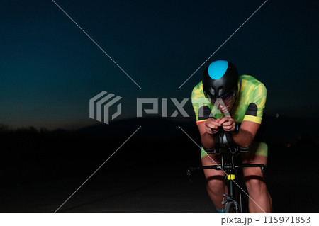A triathlete rides his bike in the darkness of night, pushing himself to prepare for a marathon. The contrast between the darkness and the light of his bike creates a sense of drama and highlights the 115971853