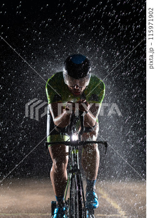 A triathlete braving the rain as he cycles through the night, preparing himself for the upcoming marathon. The blurred raindrops in the foreground and the dark, moody atmosphere in the background add A triathlete braving the rain as he cycles through the night, preparing himself for the upcoming marathon. The blurred raindrops in the foreground and the dark, moody atmosphere in the background add 115971902