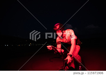 A triathlete rides his bike in the darkness of night, pushing himself to prepare for a marathon. The contrast between the darkness and the light of his bike creates a sense of drama and highlights the A triathlete rides his bike in the darkness of night, pushing himself to prepare for a marathon. The contrast between the darkness and the light of his bike creates a sense of drama and highlights the 115971906