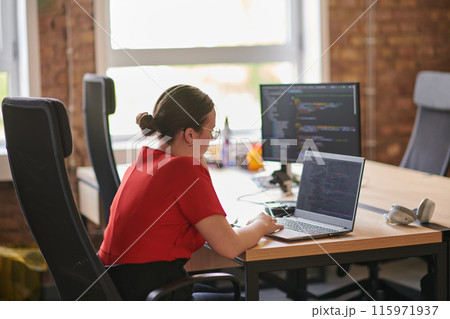 In a contemporary office setting, a young businesswoman is focused on her laptop, displaying dedication and efficiency in her work 115971937