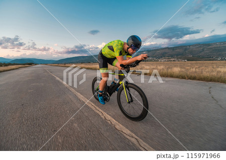 Triathlete riding his bicycle during sunset, preparing for a marathon. The warm colors of the sky provide a beautiful backdrop for his determined and focused effort. 115971966