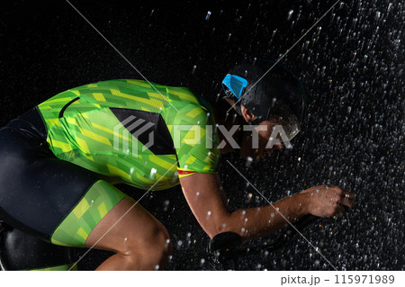 A triathlete braving the rain as he cycles through the night, preparing himself for the upcoming marathon. The blurred raindrops in the foreground and the dark, moody atmosphere in the background add 115971989