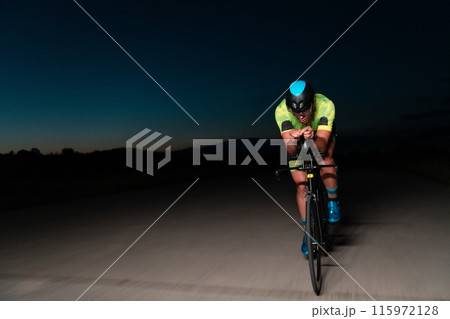 A triathlete rides his bike in the darkness of night, pushing himself to prepare for a marathon. The contrast between the darkness and the light of his bike creates a sense of drama and highlights the A triathlete rides his bike in the darkness of night, pushing himself to prepare for a marathon. The contrast between the darkness and the light of his bike creates a sense of drama and highlights the 115972128