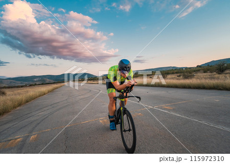 Triathlete riding his bicycle during sunset, preparing for a marathon. The warm colors of the sky provide a beautiful backdrop for his determined and focused effort. Triathlete riding his bicycle during sunset, preparing for a marathon. The warm colors of the sky provide a beautiful backdrop for his determined and focused effort. 115972310