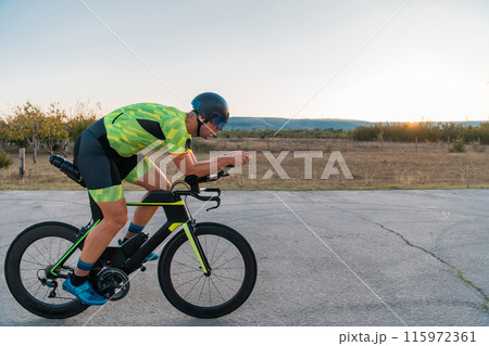Triathlete riding his bicycle during sunset, preparing for a marathon. The warm colors of the sky provide a beautiful backdrop for his determined and focused effort. Triathlete riding his bicycle during sunset, preparing for a marathon. The warm colors of the sky provide a beautiful backdrop for his determined and focused effort. 115972361