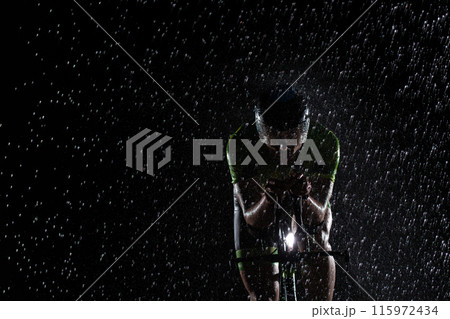 A triathlete braving the rain as he cycles through the night, preparing himself for the upcoming marathon. The blurred raindrops in the foreground and the dark, moody atmosphere in the background add A triathlete braving the rain as he cycles through the night, preparing himself for the upcoming marathon. The blurred raindrops in the foreground and the dark, moody atmosphere in the background add 115972434