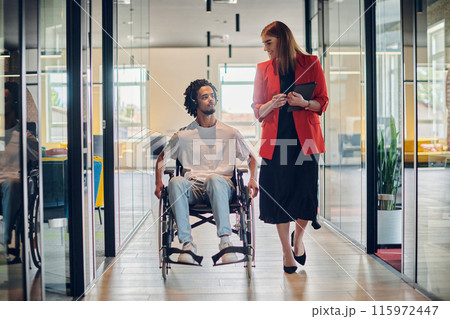 A group of young business people in a modern glass-walled office captures the essence of diversity and collaboration, while two colleagues, including an African American businessman in a wheelchair 115972447