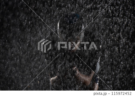 A triathlete braving the rain as he cycles through the night, preparing himself for the upcoming marathon. The blurred raindrops in the foreground and the dark, moody atmosphere in the background add 115972452