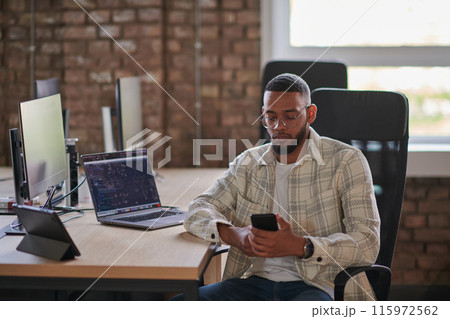 A young African American entrepreneur takes a break in a modern office, using a smartphone to browse social media, capturing a moment of digital connectivity and relaxation amidst his business 115972562