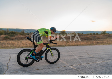 Triathlete riding his bicycle during sunset, preparing for a marathon. The warm colors of the sky provide a beautiful backdrop for his determined and focused effort. 115972592