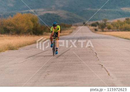 Triathlete riding his bicycle during sunset, preparing for a marathon. The warm colors of the sky provide a beautiful backdrop for his determined and focused effort. Triathlete riding his bicycle during sunset, preparing for a marathon. The warm colors of the sky provide a beautiful backdrop for his determined and focused effort. 115972623