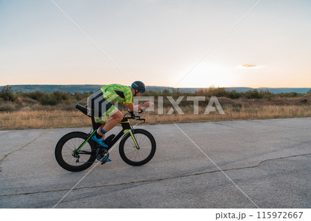 Triathlete riding his bicycle during sunset, preparing for a marathon. The warm colors of the sky provide a beautiful backdrop for his determined and focused effort. 115972667