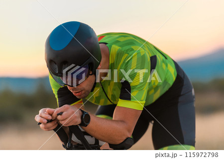 Close up photo of triathlete riding his bicycle during sunset, preparing for a marathon. The warm colors of the sky provide a beautiful backdrop for his determined and focused effort. 115972798