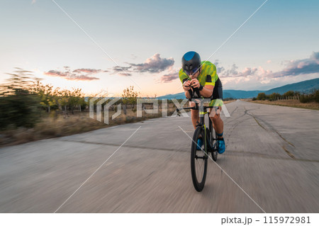 Triathlete riding his bicycle during sunset, preparing for a marathon. The warm colors of the sky provide a beautiful backdrop for his determined and focused effort. Triathlete riding his bicycle during sunset, preparing for a marathon. The warm colors of the sky provide a beautiful backdrop for his determined and focused effort. 115972981