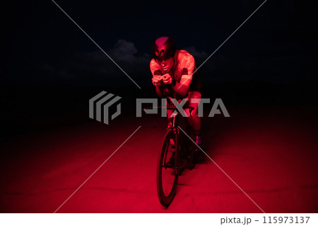 A triathlete rides his bike in the darkness of night, pushing himself to prepare for a marathon. The contrast between the darkness and the light of his bike creates a sense of drama and highlights the A triathlete rides his bike in the darkness of night, pushing himself to prepare for a marathon. The contrast between the darkness and the light of his bike creates a sense of drama and highlights the 115973137