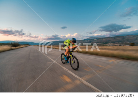 Triathlete riding his bicycle during sunset, preparing for a marathon. The warm colors of the sky provide a beautiful backdrop for his determined and focused effort. 115973207