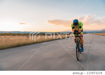 Triathlete riding his bicycle during sunset, preparing for a marathon. The warm colors of the sky provide a beautiful backdrop for his determined and focused effort. 115973234