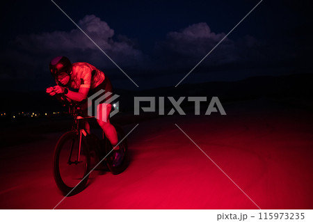 A triathlete rides his bike in the darkness of night, pushing himself to prepare for a marathon. The contrast between the darkness and the light of his bike creates a sense of drama and highlights the 115973235