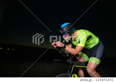 A triathlete rides his bike in the darkness of night, pushing himself to prepare for a marathon. The contrast between the darkness and the light of his bike creates a sense of drama and highlights the 115973241