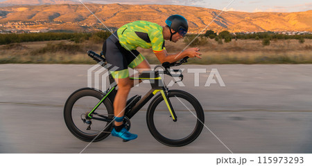 Triathlete riding his bicycle during sunset, preparing for a marathon. The warm colors of the sky provide a beautiful backdrop for his determined and focused effort. Triathlete riding his bicycle during sunset, preparing for a marathon. The warm colors of the sky provide a beautiful backdrop for his determined and focused effort. 115973293
