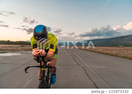 Triathlete riding his bicycle during sunset, preparing for a marathon. The warm colors of the sky provide a beautiful backdrop for his determined and focused effort. Triathlete riding his bicycle during sunset, preparing for a marathon. The warm colors of the sky provide a beautiful backdrop for his determined and focused effort. 115973332