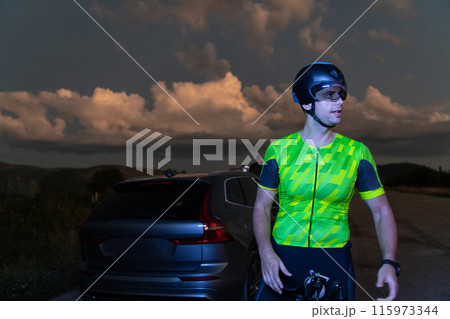 A triathlete resting on the road after a tough bike ride in the dark night, leaning on his bike in complete exhaustion  115973344