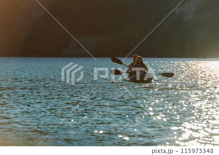 A young couple enjoying an idyllic kayak ride in the middle of a beautiful river surrounded by forest greenery in sunset time  115973348