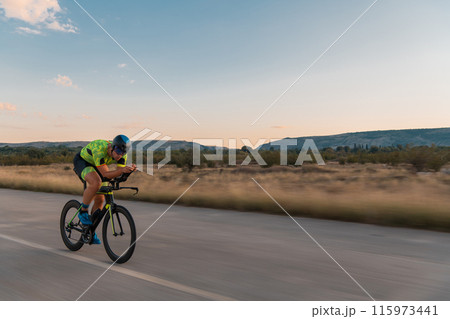 Triathlete riding his bicycle during sunset, preparing for a marathon. The warm colors of the sky provide a beautiful backdrop for his determined and focused effort. Triathlete riding his bicycle during sunset, preparing for a marathon. The warm colors of the sky provide a beautiful backdrop for his determined and focused effort. 115973441