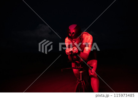 A triathlete rides his bike in the darkness of night, pushing himself to prepare for a marathon. The contrast between the darkness and the light of his bike creates a sense of drama and highlights the A triathlete rides his bike in the darkness of night, pushing himself to prepare for a marathon. The contrast between the darkness and the light of his bike creates a sense of drama and highlights the 115973485