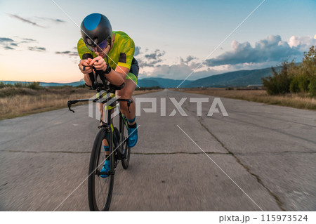 Triathlete riding his bicycle during sunset, preparing for a marathon. The warm colors of the sky provide a beautiful backdrop for his determined and focused effort. 115973524