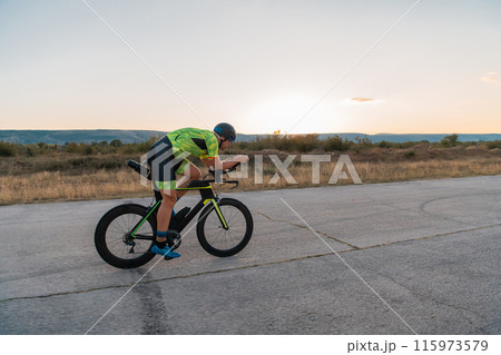 Triathlete riding his bicycle during sunset, preparing for a marathon. The warm colors of the sky provide a beautiful backdrop for his determined and focused effort. 115973579