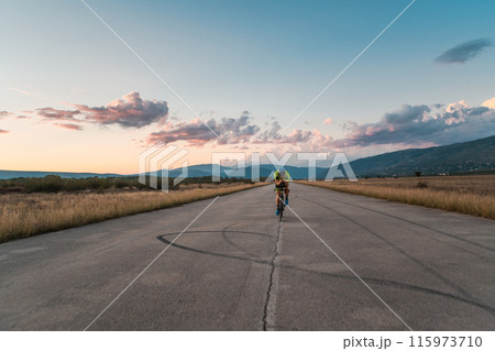 Triathlete riding his bicycle during sunset, preparing for a marathon. The warm colors of the sky provide a beautiful backdrop for his determined and focused effort. 115973710