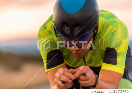 Close up photo of triathlete riding his bicycle during sunset, preparing for a marathon. The warm colors of the sky provide a beautiful backdrop for his determined and focused effort. Close up photo of triathlete riding his bicycle during sunset, preparing for a marathon. The warm colors of the sky provide a beautiful backdrop for his determined and focused effort. 115973746