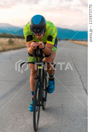 Triathlete riding his bicycle during sunset, preparing for a marathon. The warm colors of the sky provide a beautiful backdrop for his determined and focused effort. Triathlete riding his bicycle during sunset, preparing for a marathon. The warm colors of the sky provide a beautiful backdrop for his determined and focused effort. 115973770