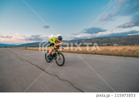 Triathlete riding his bicycle during sunset, preparing for a marathon. The warm colors of the sky provide a beautiful backdrop for his determined and focused effort. Triathlete riding his bicycle during sunset, preparing for a marathon. The warm colors of the sky provide a beautiful backdrop for his determined and focused effort. 115973853