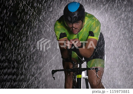 A triathlete braving the rain as he cycles through the night, preparing himself for the upcoming marathon. The blurred raindrops in the foreground and the dark, moody atmosphere in the background add A triathlete braving the rain as he cycles through the night, preparing himself for the upcoming marathon. The blurred raindrops in the foreground and the dark, moody atmosphere in the background add 115973881