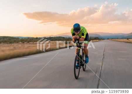 Triathlete riding his bicycle during sunset, preparing for a marathon. The warm colors of the sky provide a beautiful backdrop for his determined and focused effort. 115973930