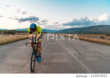 Triathlete riding his bicycle during sunset, preparing for a marathon. The warm colors of the sky provide a beautiful backdrop for his determined and focused effort. 115973931