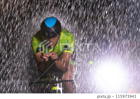 A triathlete braving the rain as he cycles through the night, preparing himself for the upcoming marathon. The blurred raindrops in the foreground and the dark, moody atmosphere in the background add 115973941