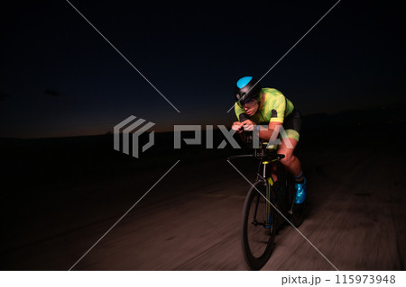 A triathlete rides his bike in the darkness of night, pushing himself to prepare for a marathon. The contrast between the darkness and the light of his bike creates a sense of drama and highlights the 115973948