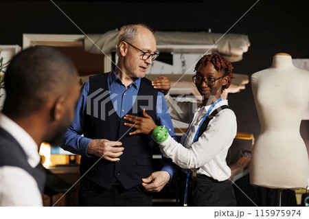 African american tailoring assistant happy with the way bespoke sartorial piece fits elderly customer, waiting for approval from master couturier. Client trying on premium quality suit vest 115975974