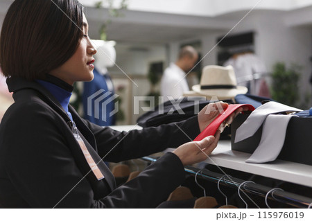 Clothing store asian woman employee showcasing accessory for sale on display shelf. Seller holding red tie while examining merchandise stock in shopping department center boutique 115976019