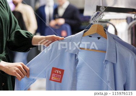 Close up of unrecognizable woman shopper looking at clothes with red discount tag while shopping in outlet store during Black Friday. Female customer holding shirt making purchases in sale season 115976115
