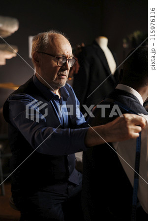 Apprentice suitmaker eager to help master couturier with sartorial measurements for upcoming bespoke fashion collection, close up. Skilled tailor checking handmade suit fitting 115976116