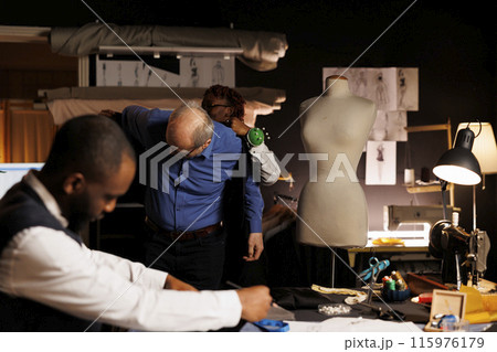 Needleworker helping senior client try on unfinished suit garment while professional fashion designer picks best adjustments to be made. Couturiers helping elderly costumer with custom made attire Needleworker helping senior client try on unfinished suit garment while professional fashion designer picks best adjustments to be made. Couturiers helping elderly costumer with custom made attire 115976179