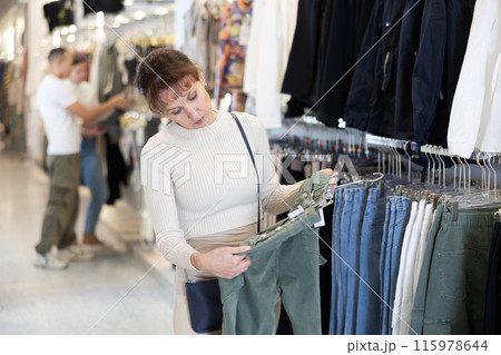 Woman buyer in store has removed hanger with jeans from window and is considering product. Woman buyer in store has removed hanger with jeans from window and is considering product. 115978644